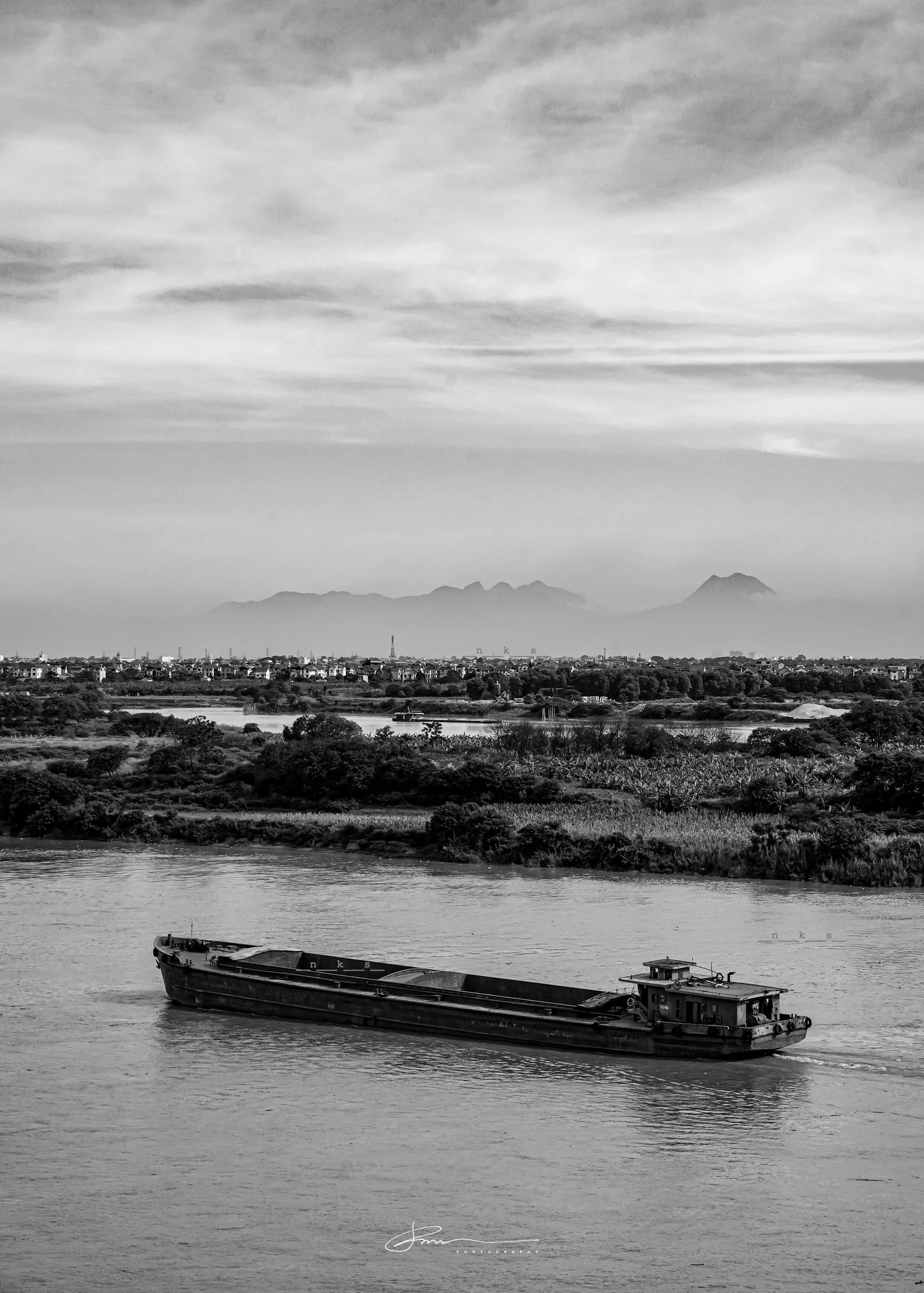 Một sà lan chở hàng di chuyển trên sông với dãy núi mờ ảo phía xa gần Tam Đảo, ảnh đen trắng.